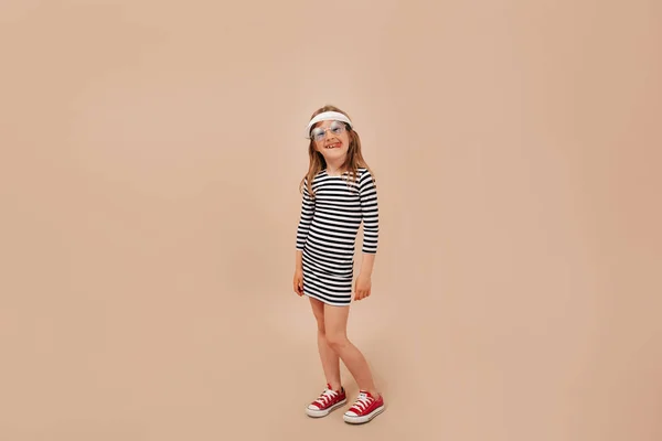 Full-lenght foto gadis kecil yang lucu menawan mengenakan gaun, topi dan sepatu berpose di kamera di atas latar beige — Stok Foto
