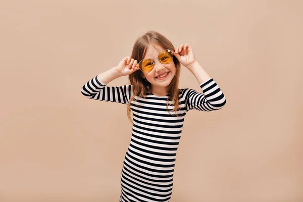 Divertido feliz niña de 6 años en vestido despojado con gafas redondas de color naranja mirando hacia otro lado con encantadora sonrisa sobre fondo beige — Foto de Stock