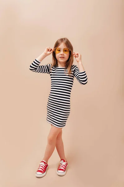 Foto de larga duración de la niña feliz con vestido despojado y gafas redondas con estilo tiene diversión sobre fondo beige aislado — Foto de Stock