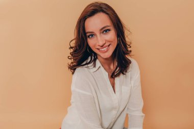 Kıvırcık koyu saçlı mutlu inanılmaz kadın kameraya bakıyor ve beyaz gömlek giyen bej arka plan üzerinde gülümseyen