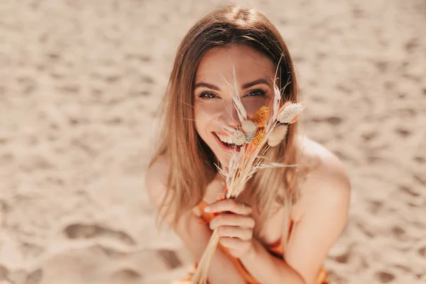 Vicino all'esterno ritratto di affascinante bella signora che copre il viso con fiori estivi e seduto sulla spiaggia di sabbia alla luce del sole — Foto Stock
