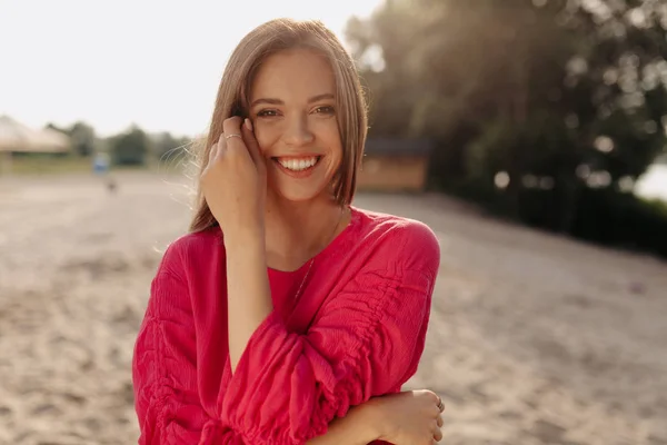 Wanita cantik dengan senyum lembut menatap kamera di latar belakang pantai berpasir di bawah sinar matahari — Stok Foto
