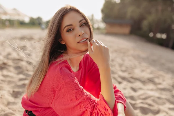 Wanita Kaukasia cantik dengan rambut panjang mengenakan pakaian musim panas merah muda duduk di pantai berpasir di bawah sinar matahari dan melihat kamera — Stok Foto