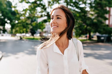 Güneşli yaz parkında yürüyen beyaz elbise giyen koyu saçlı Avrupalı kadının açık hava yakın portresi