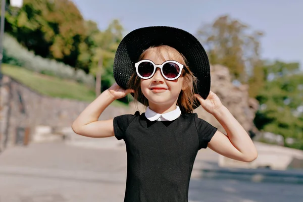 Close up tiro ao ar livre de menina encantadora vestindo vestido preto e chapéu segurando chapéu e andando à luz do sol — Fotografia de Stock