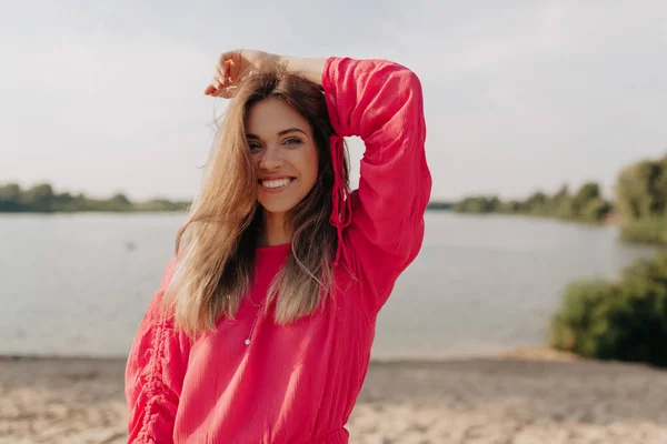 Feliz hermosa mujer caucásica sonriendo durante la sesión de fotos al aire libre y sosteniendo la mano en la cabeza en el fondo de la playa de verano — Foto de Stock