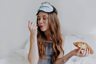 Uzun dalgalı saçlı, yatakta uyku maskesi takan, elinde kruvasan ve kapalı gözlü fındıklı bir kız. 