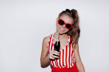 Çekici bir genç kadının portresi. Kırmızı güneş gözlüğü takıyor ve tişörtünü çıkarıp kola içiyor..