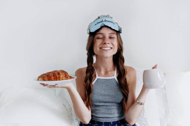 Saçları örgülü, kafasında uyku maskesiyle yatakta oturan tatmin olmuş bir kız. 