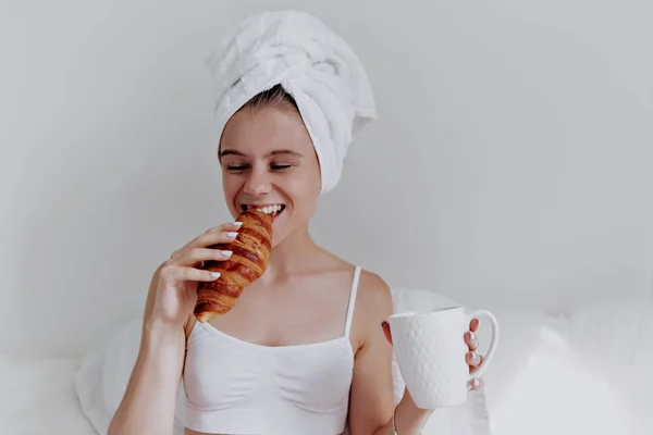 Potret wanita muda yang cantik minum kopi dan menggigit croissant dengan handuk di kepala di tempat tidur putih — Stok Foto