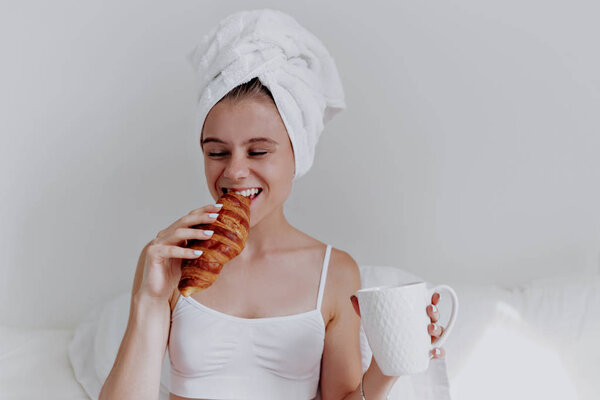 Portrait of beautiful young woman drinking coffee and biting croissant with towel on the head on white bed