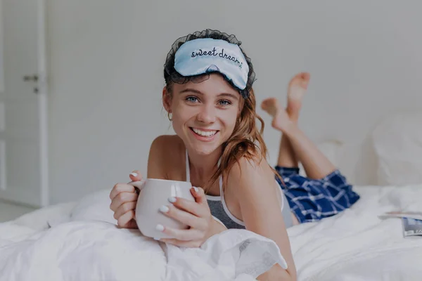 Chica feliz usando ropa de dormir y máscara de dormir acostado en la cama con una taza y sonriendo —  Fotos de Stock
