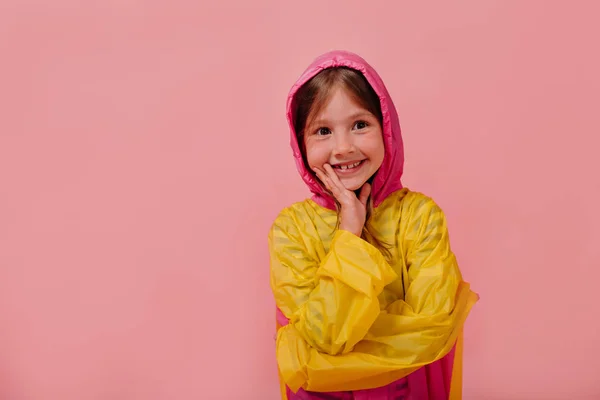 Sonriente chica feliz usando impermeable brillante sonriendo sobre el fondo rosa aislado y sosteniendo la mano cerca de la cara — Foto de Stock