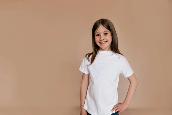 Senyum yakin gadis kecil di t-shirt putih melihat kamera di latar belakang terisolasi. Potret dalam ruangan gadis cantik yang cantik Stok Foto Bebas Royalti