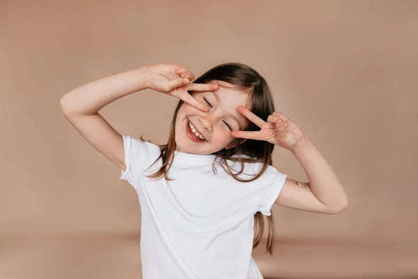 Engraçado rindo pequena garota europeia posando no estúdio sobre fundo bege com sorriso e olhos fechados e mostrando sinal de paz — Fotografia de Stock