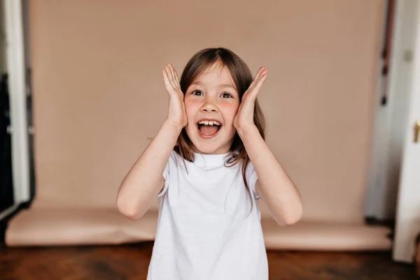 Adorável menina emocional vestindo t-shirt branca se divertindo, sorrindo, de mãos dadas perto do rosto sobre fundo bege . — Fotografia de Stock