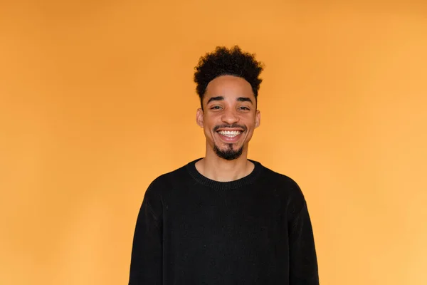 Retrato de estudio de un hombre afroamericano con un jersey negro sonriendo sobre fondo naranja. Retrato de un hombre alegre de piel oscura. Vista frontal . —  Fotos de Stock