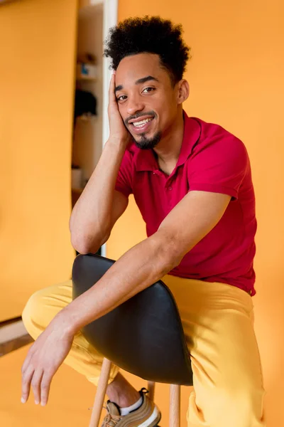 Retrato interior de un hombre afro americano feliz con camiseta roja y pantalones amarillos sentados en la silla en el estudio sobre fondo naranja. Estudio disparo de hombre guapo —  Fotos de Stock