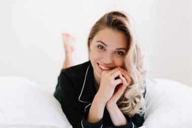 Sarı saçlı, takım elbiseli, beyaz yatakta yatıp kameraya poz veren mutlu bir kadının portresini yakından çek.. 