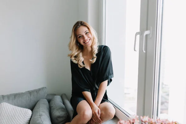 Wanita muda yang menawan dengan senyum yang indah mengenakan jubah mandi hitam duduk di dekat jendela di rumah. Selamat pagi di rumah. — Stok Foto