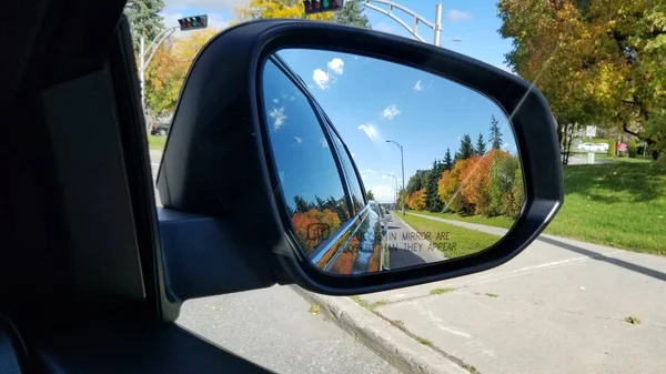 Reflection of asphalt highway road with cars and trees at the car side mirror. The road in car mirror with blue sky and green, orange trees in the background.