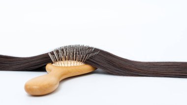 Parlak beyaz bir arkaplanda parlak kahverengi saçlara yerleştirilmiş ahşap saç fırçası, saç bakım araçları ve güzellik meraklıları için bakım uygulamaları vurgulanıyor