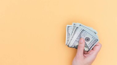 Close-up of a hand holding a fan of U.S. dollar banknotes, illustrating financial wealth, success, savings, and money handling. Isolated on a light yellow background.