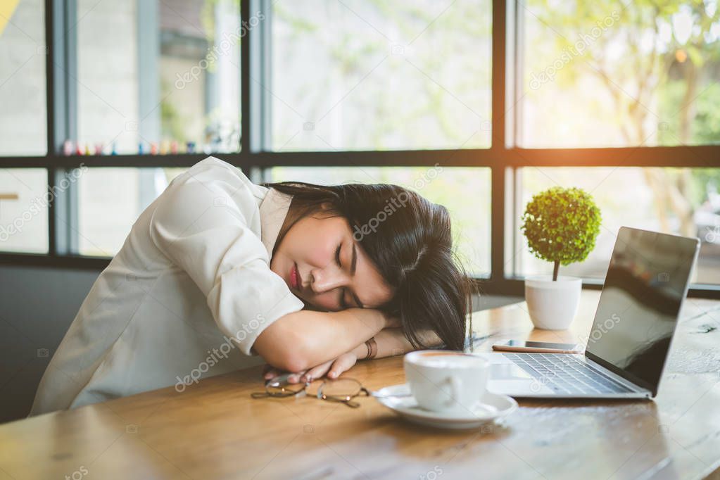 Freelancer asian businesswoman tired after working coffee shop her sleeping on workplace table near windows at evening with digital laptop computer and coffe break.