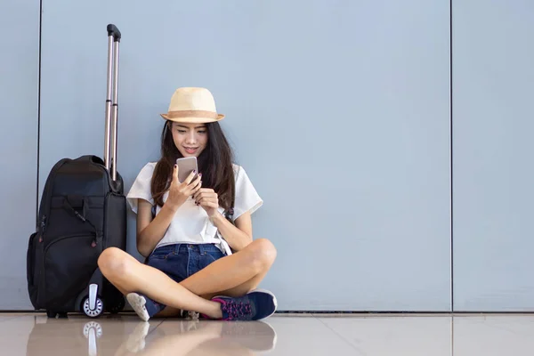 Asian woman teenager using smartphone at airport terminal sitting with luggage suitcase and backpack for travel in vacation summer relaxing waiting flight transport online check in or booking ticket