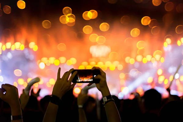 Crowd holding smartphone at concert stage lights and people fan audience silhouette raising hands in the music festival rear view with spotlight glowing effect bokeh