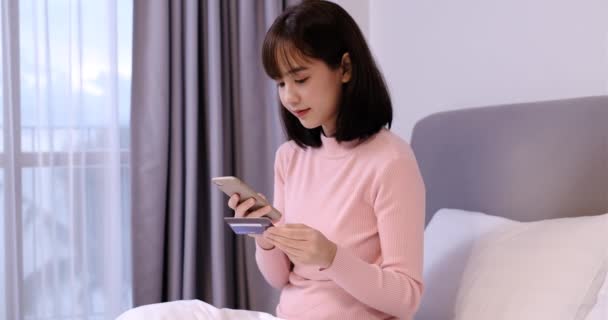 若いアジアの女性は スマートフォンのオンラインアカウントをチェックし クレジットカードを保持し 快適なベッドに横たわっている間にオンラインショッピングを行います 素晴らしい若い女性は新しい購入を喜んでいます — ストック動画