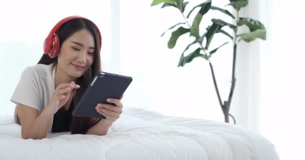 デジタルタブレットの笑顔でヘッドフォンを着用し 音楽を聴くことを楽しむ若いアジアの女性 美しい女性が朝にベッドの上に横になり 誠実に笑って — ストック動画