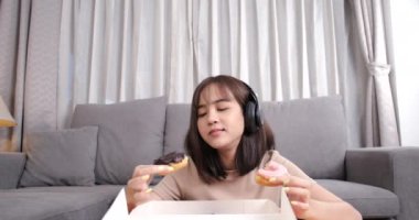 Genç Asyalı bir kadın kanepenin üzerinde abur cubur yiyip oturma odasında müzik dinliyor, Fast Food servis hizmetleri güvende ve Coronavirus 'un sosyal uzaklık konseptiyle yayılmasını engelliyor..
