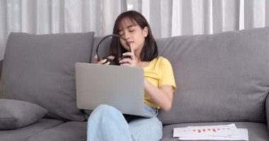 Genç Asyalı kadın kulaklık takıyor arkadaşlarıyla görüntülü konuşma yapıyor oturma odasındaki koltukta otururken dizüstü bilgisayar kullanıyor, internetteki sosyal mesafeli konuşma için video görüşmesi yapıyor..