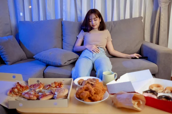Young asian woman takeaway eating full junk food sitting on sofa and watch television in living room at home Fast food delivery service, She is already tried to sleep.