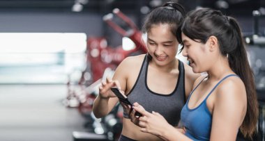 Spor salonundaki antrenmanlarından sonra gevşeyen ve spor salonunda gülümseyen iki genç Asyalı kadın cep telefonlarını kontrol edip fotokopi alanındaki sağlıklı yaşam tarzlarını izliyorlar..