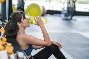 Sportif genç Asyalı kadın spor salonunda başarılı bir şekilde çalıştıktan sonra elinde bir şişe protein içeceği, yerde oturan sağlıklı bir vücut geliştirme sporcu kasları konsepti..