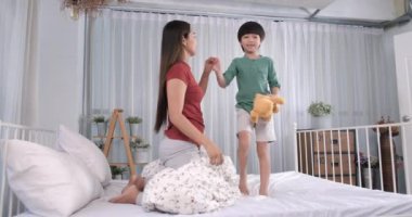 Asyalı bir anne, oğlunun elini tutarken yatakta arkasını dönüyor. Anne ve küçük çocuk, evde yatakta masal anlatırken eğleniyorlar. Mutlu çocukluk, özen, sevgi.