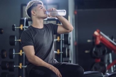 Spor salonunda tişörtlü su içen yakışıklı sporcu, egzersiz yaptıktan sonra dinleniyor. Alıştırma sonrası çekici atletik adam rahatlama konsepti.