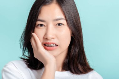Diş teli takan genç Asyalı kadın mutsuz, kendine güveni yok. Mavi arka planda, izole edilmiş stüdyo çekimlerinde sağlıklı..