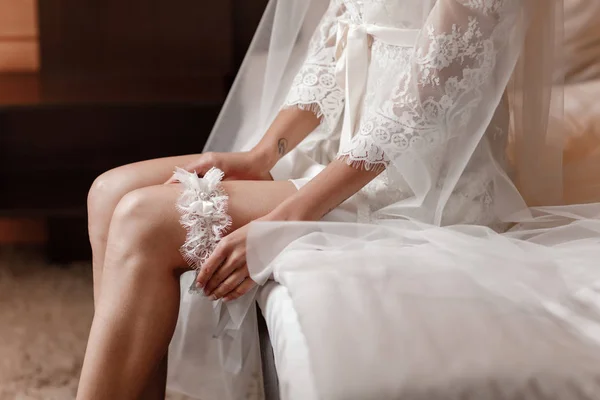 女性の脚にガーターを着用します。花嫁は、ホテルの部屋で手で失うまでガーターを保持しています。朝の準備結婚式のコンセプト. — ストック写真