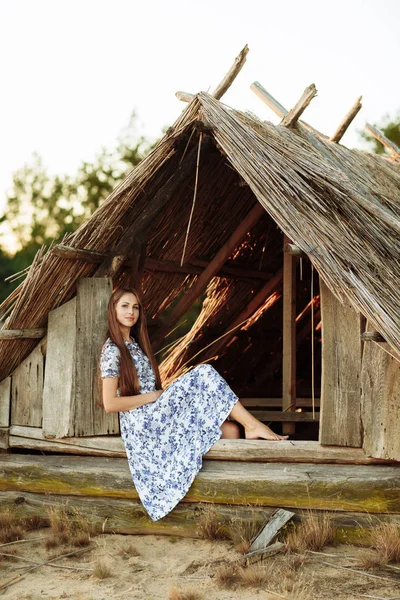 Porträt eines schönen Mädchens vor einem Holzhaus. Mädchen auf dem Hintergrund eines alten Holzhauses geworfen. Mädchen im weißen Kleid mit blauen Stickblumen. — Stockfoto