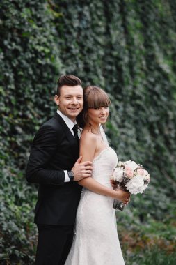 Romantik mutlu çift yeni evliler, gelin ve damat standları ve pembe ve mor çiçek ve yeşillik buket tutan, bahçede kurdele ile yeşillik. Doğa üzerine düğün töreni.