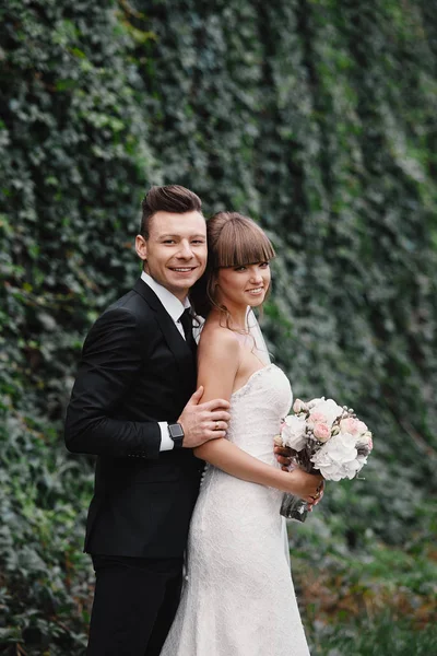 Romantisches glückliches Paar, Braut und Bräutigam steht und hält einen Strauß aus rosa und lila Blumen und Grüntönen, Grün mit Schleife im Garten. Hochzeitsfeier in der Natur. — Stockfoto
