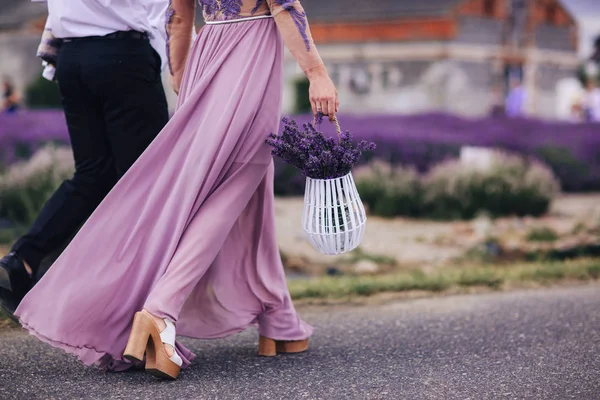 Hermosa mujer joven en vestido azul sostiene ramo de flores de lavanda en cesta mientras camina al aire libre a través del campo de trigo al atardecer en verano. Provenza, Francia. Imagen tonificada con espacio de copia . —  Fotos de Stock