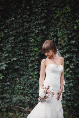 Young beautiful bride in an elegant dress is standing on the field near the forest and holding bouquet of pink flowers and greens with ribbon at nature. Outdoors. After the wedding ceremony.