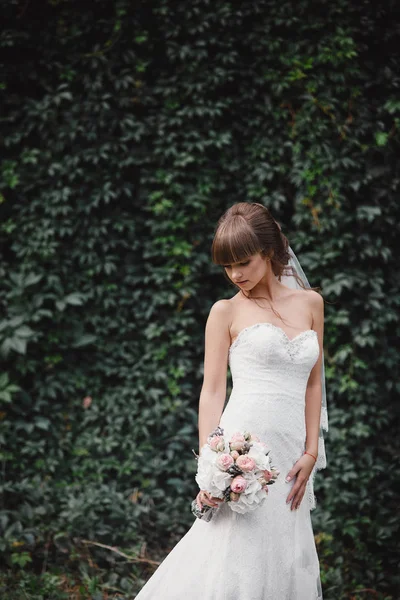 Jovem noiva bonita em um vestido elegante está de pé no campo perto da floresta e segurando buquê de flores rosa e verdes com fita na natureza. Ao ar livre. Após a cerimônia de casamento . — Fotografia de Stock