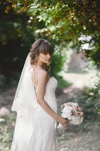 Νεαρή όμορφη νύφη σε ένα κομψό φόρεμα στέκεται στο γήπεδο κοντά στο δάσος και κρατώντας μπουκέτο από ροζ λουλούδια και χόρτα με κορδέλα στη φύση. Έξω. Μετά την τελετή του γάμου. — Φωτογραφία Αρχείου