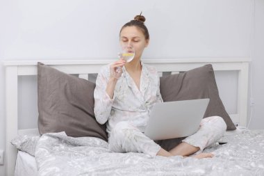 Genç bir kadın laptopunu yatakta dinlenirken ve şarap içerken kullanıyor. Evde kalmak için. Çevrimiçi çalışma. Uzak çalışma