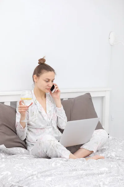 年轻女人在床上休息和喝酒的时候用她的笔记本电脑 呆在家里在线学习 远程工作 — 图库照片